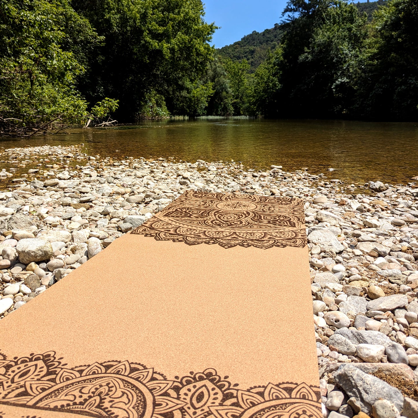 The ZenMotion Cosmic Dance Mandala cork yoga mat laid on a river bank.